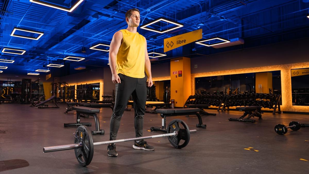 Hombre de pie en un gimnasio Smart Fit con luces azules y amarillas, usando camiseta sin mangas amarilla y pantalones negros, frente a una barra con pesas en el area de peso libre.