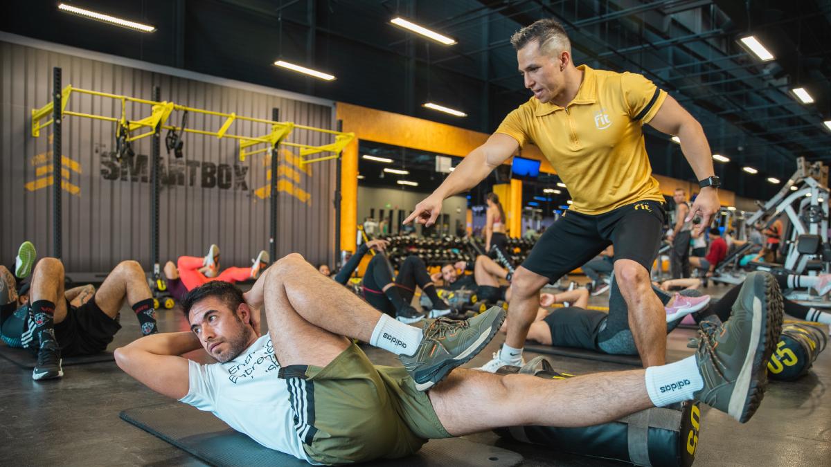 hombre recostado en el suelo con la cabeza levantada y los manos entelazadas en la nuca. una pierna estirada y la otra flexionada. profesor del gimnasio smart fit a un costado