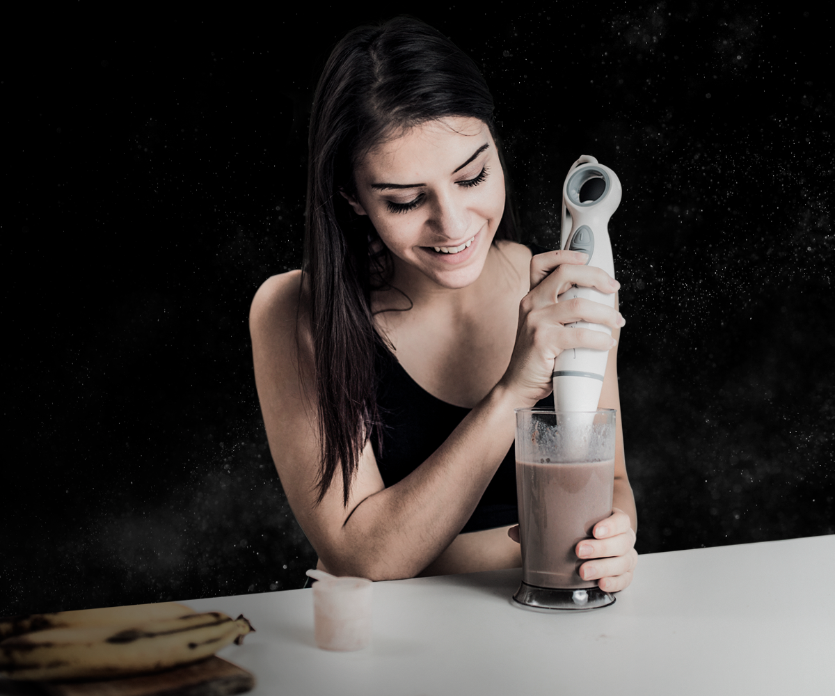 mujer con una batidora realizando un batido en un vaso. A un lado un scoop de suplementos para subir de peso y un par de plátanos
