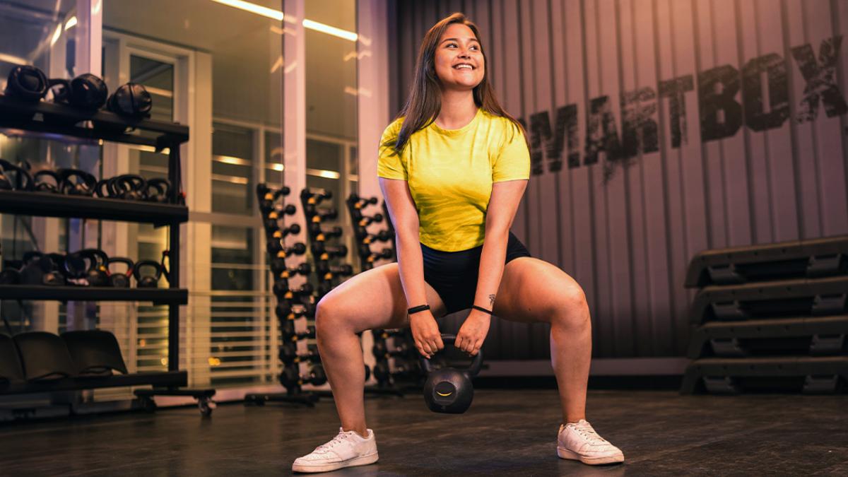 mujer de pie con las piernas separada y ligeramente felxionadas, en posición de sentadilla, con una pesa rusa sostenida con las dos manos a la altura de las rodillas