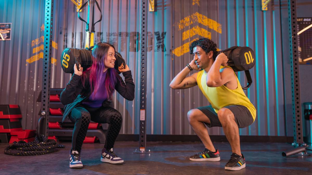 hombre y mujer de perfil con un costal de peso en la espalda realizando una sentadilla sumo