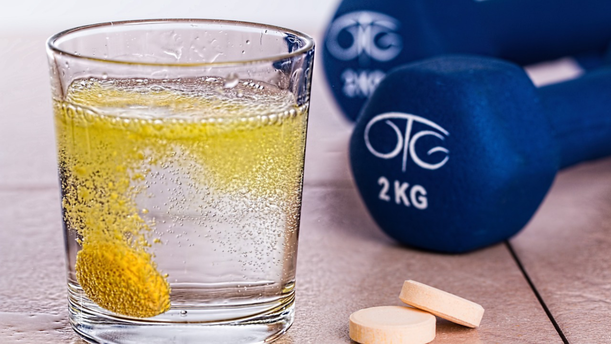 vaso con agua y una vitamina efervescente, aun lado dos tabletas y dos mancuernas