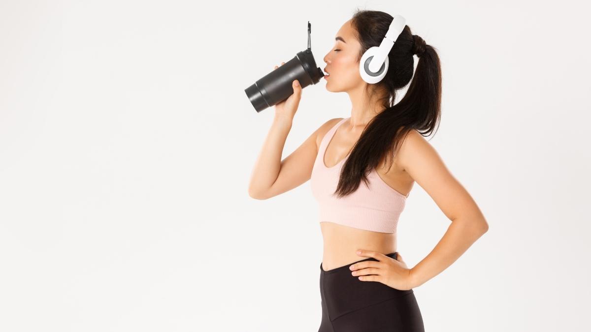 mujer de perfil, con outfit deportivo y audifonos en la cabeza, tomando una malteada de proteina en un shaker.