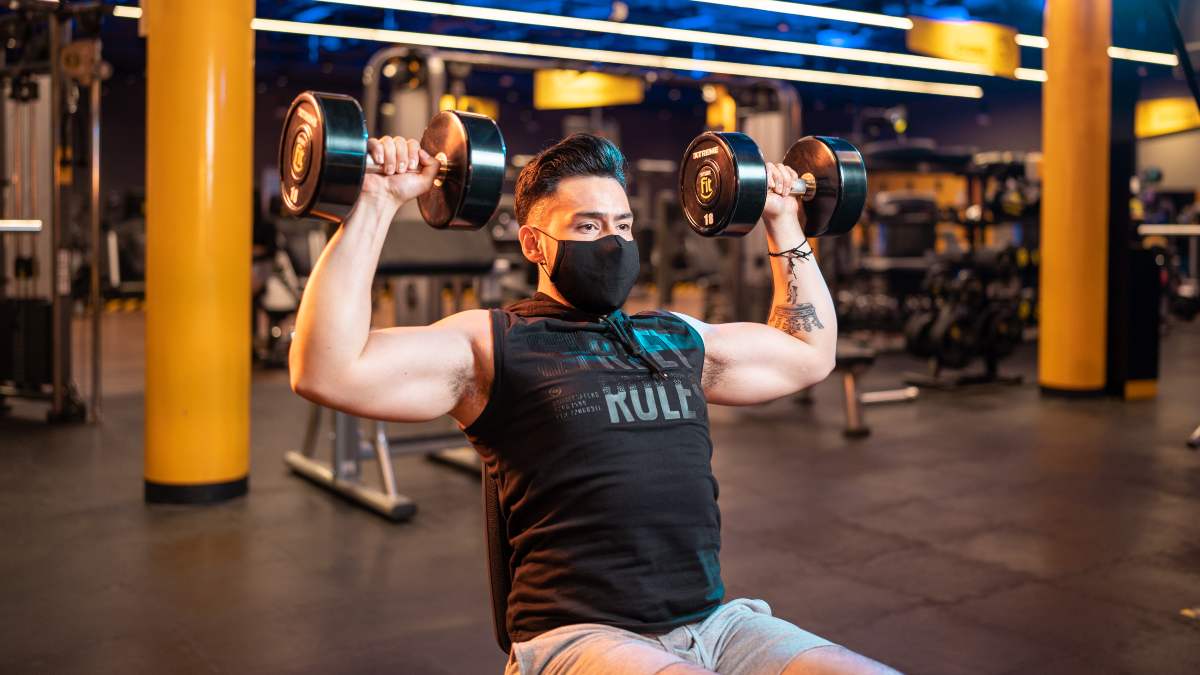 hombre en un gimnasio realizando un press militar con mancuernas sentado en un banco