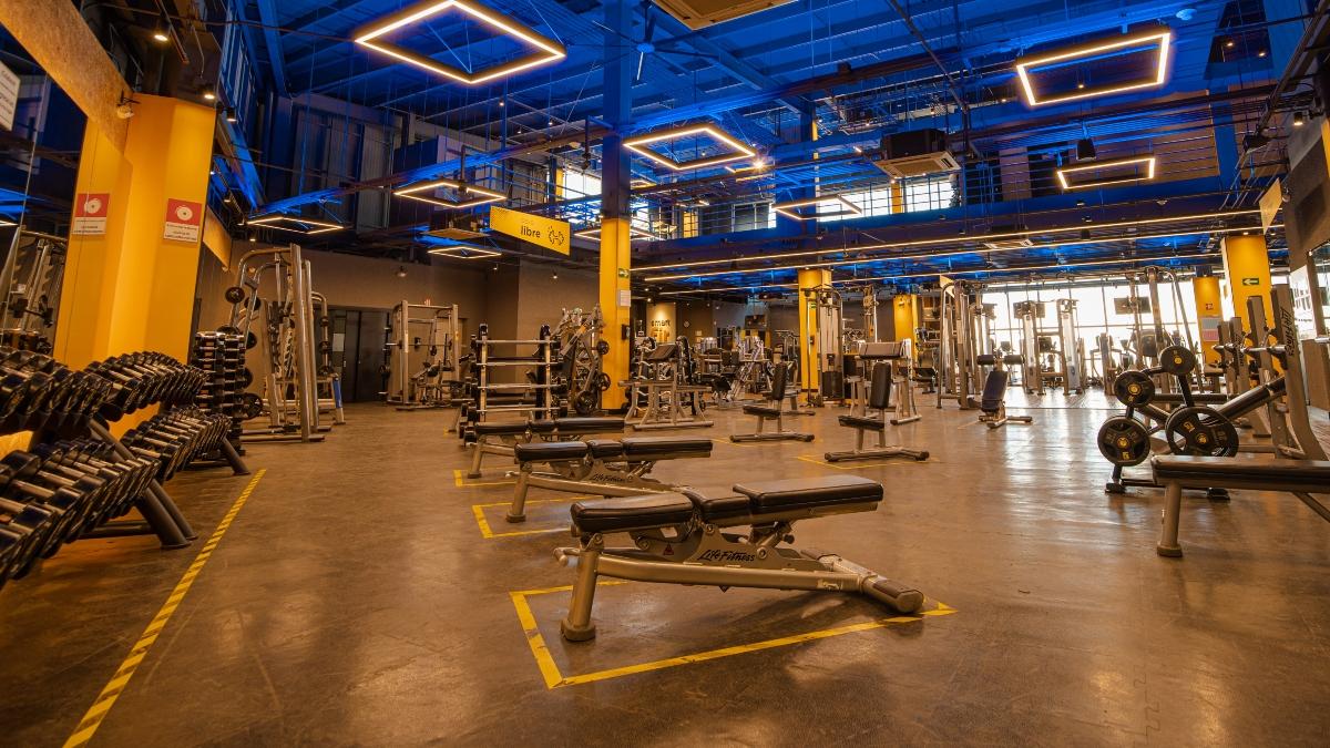 Vista amplia de un gimnasio Smart Fit con una gran variedad de equipos de entrenamiento, incluyendo pesas libres, maquinas de ejercicio y bancos de entrenamiento. La iluminacion azul y amarilla resalta el ambiente espacioso y bien organizado