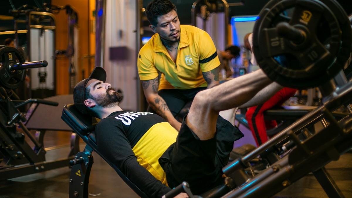 hombre sentado en una prensa del gym realizando un empuje con las piernas. A un costado un profesor del gimnasio supervisando el ejercicio