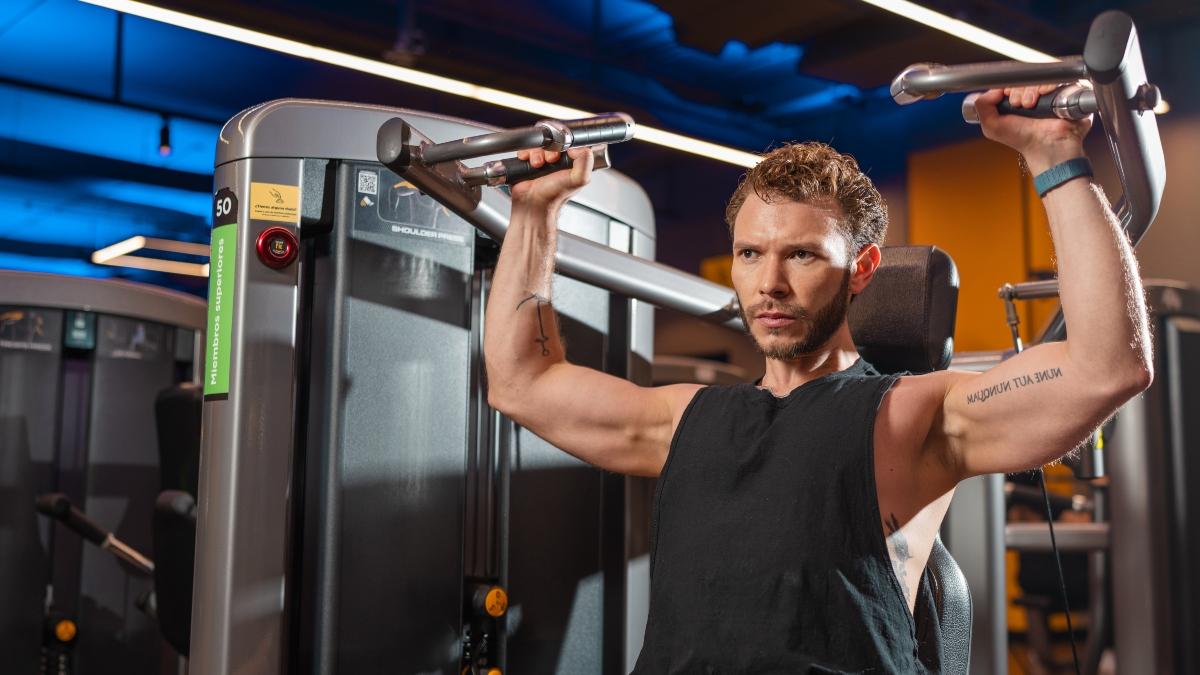 Un hombre con barba y camiseta sin mangas usa una maquina de press de hombros en un gimnasio Smart Fit