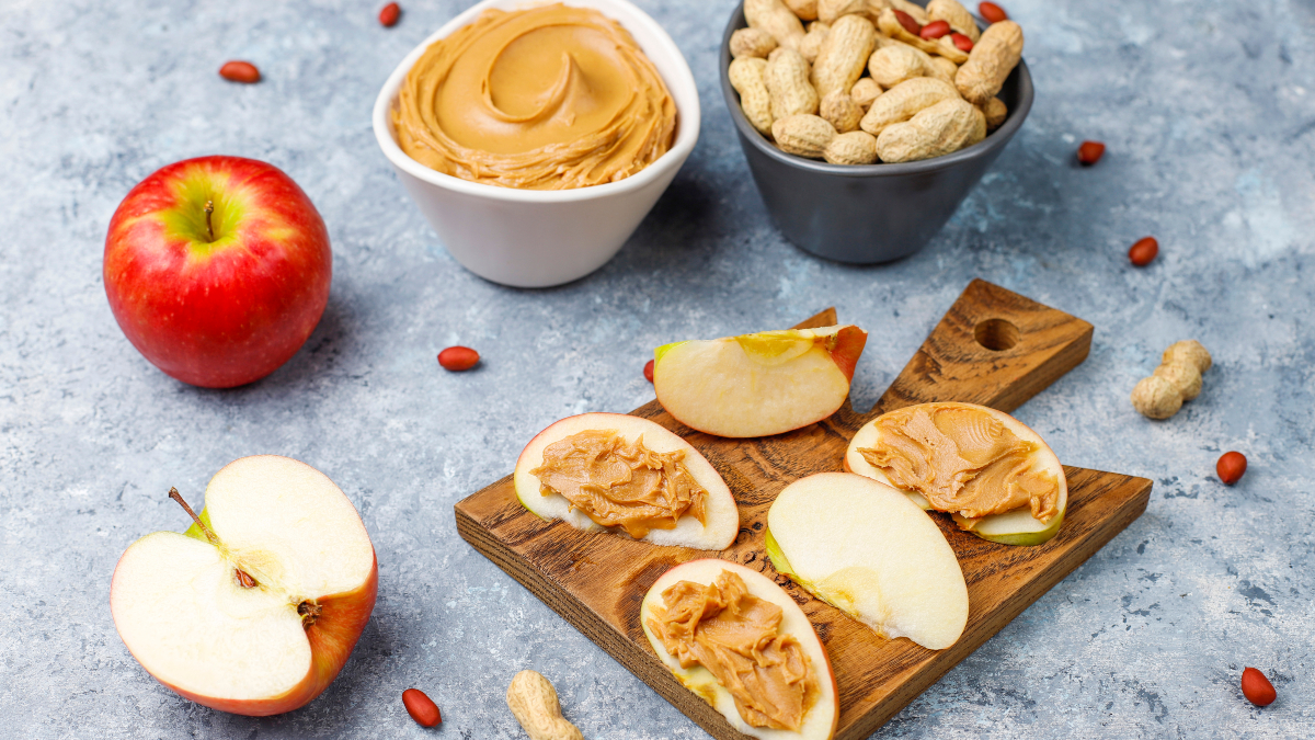 Mesa con manzanas completas y rebanadas con crema de cacahuate untada. Tazón con cacahuates con cáscara y tazón con crema de cacahuate.