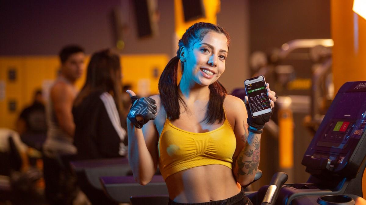torso de una mujer de frente sosteniendo en una mano un celular con una aplicación para hacer ejercicio en la pantalla. pulgar arriba con la otra mano y una sonrisa en el rostro