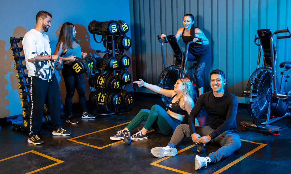 Tres mujeres y dos hombres platicando y sonriendo mientras realizan ejercicio en un area del gimnasio Smart Fit en Culiacán