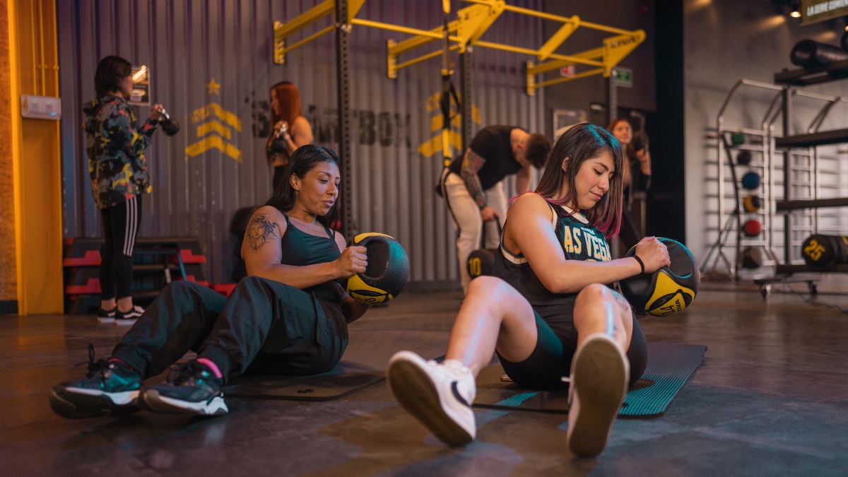 Dos mujeres sentadas sobre un tapete realizando un giro ruso con una balón de peso en las manos. En el fondo otros usuarios realizando ejercicios en el area Smart Box