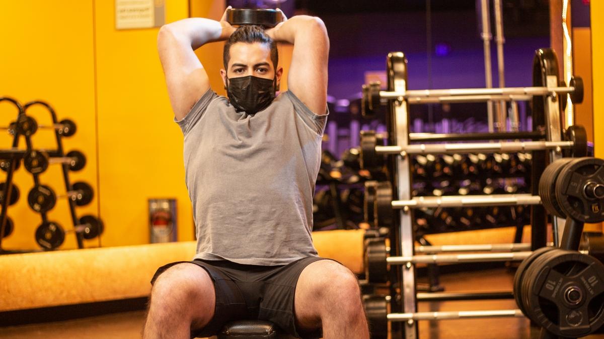 hombre sentado en un banco en el gimnasio, con una mancuerna en las manos realizando una copa a dos manos en su fase concéntrica