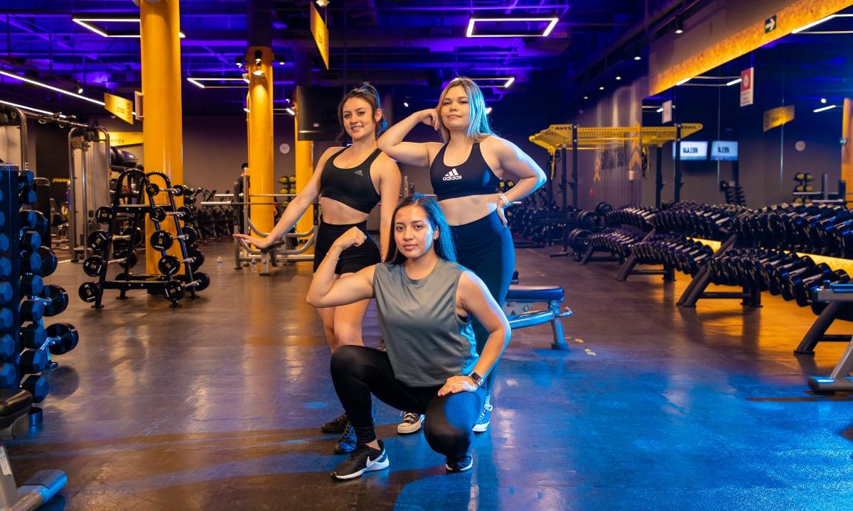 Una mujer en cuclillas y dos más de pie, mostrando sus biceps. Posando en un gimnasio de Smart Fit.