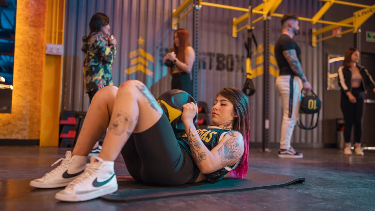 mujer recostada en el suelo con las rodillas flexionadas y un balón medicinal a la altura del abdomen sostenido con las dos manos. Está realizando un ejercicio; giro ruso