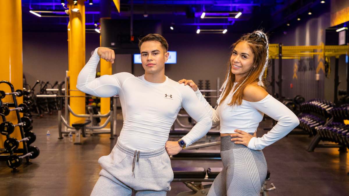 hombre parado de frente con el brazo felxionado, mostrando su bíceps. A un lado, una mujer parada de perfil tocando el hombro del hombre. Al fondo están los aparatos del gimnasio Smart Fit