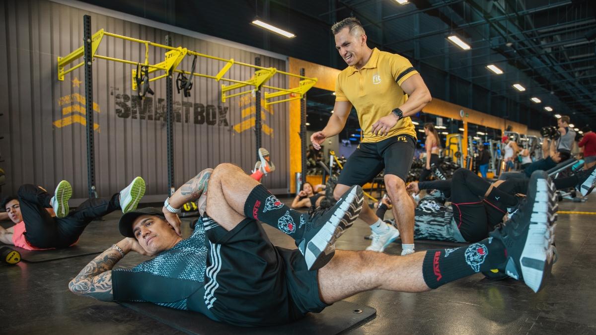 hombre recostado en el suelo encongiendo una pierna para hacer un ejercicio de abdomen. Entrenador a un lado alentándolo.