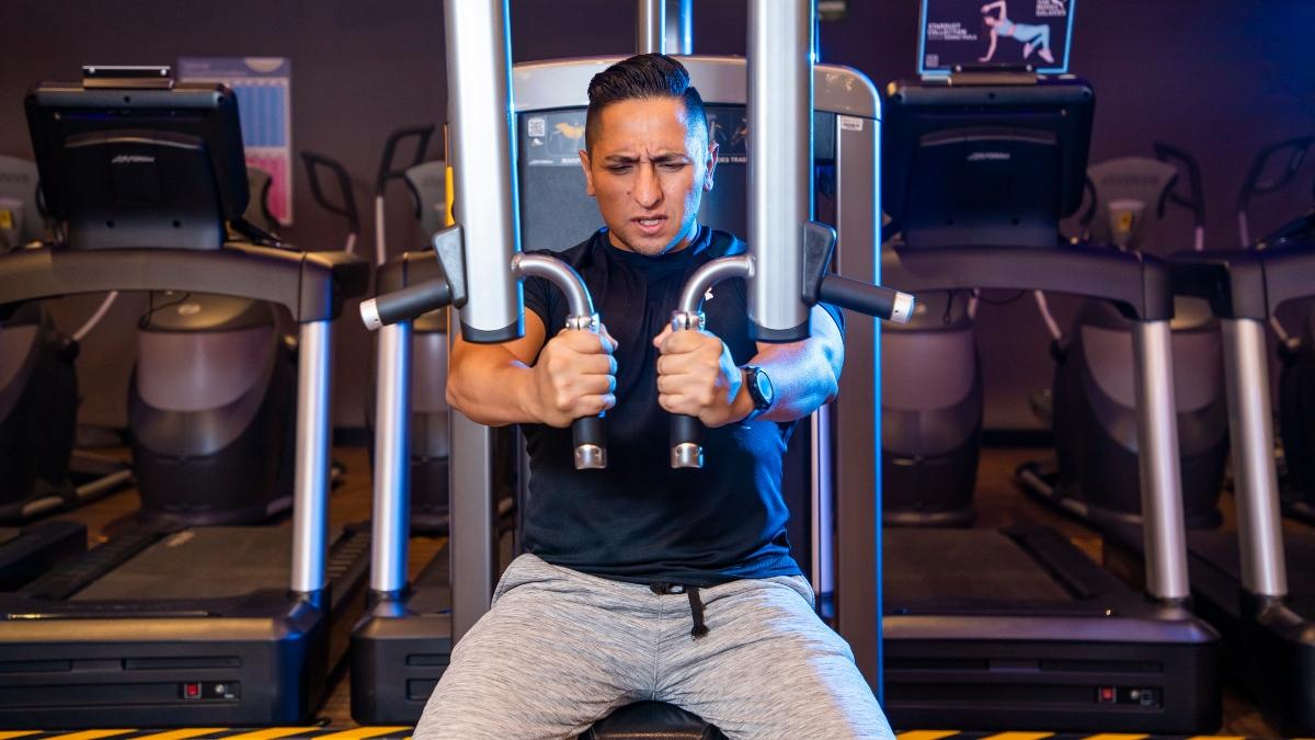 hombre sentado en una máquina del gimnasio, sosteniendo las agarraderas del aparato al frente, concentrado en el ejercicio.