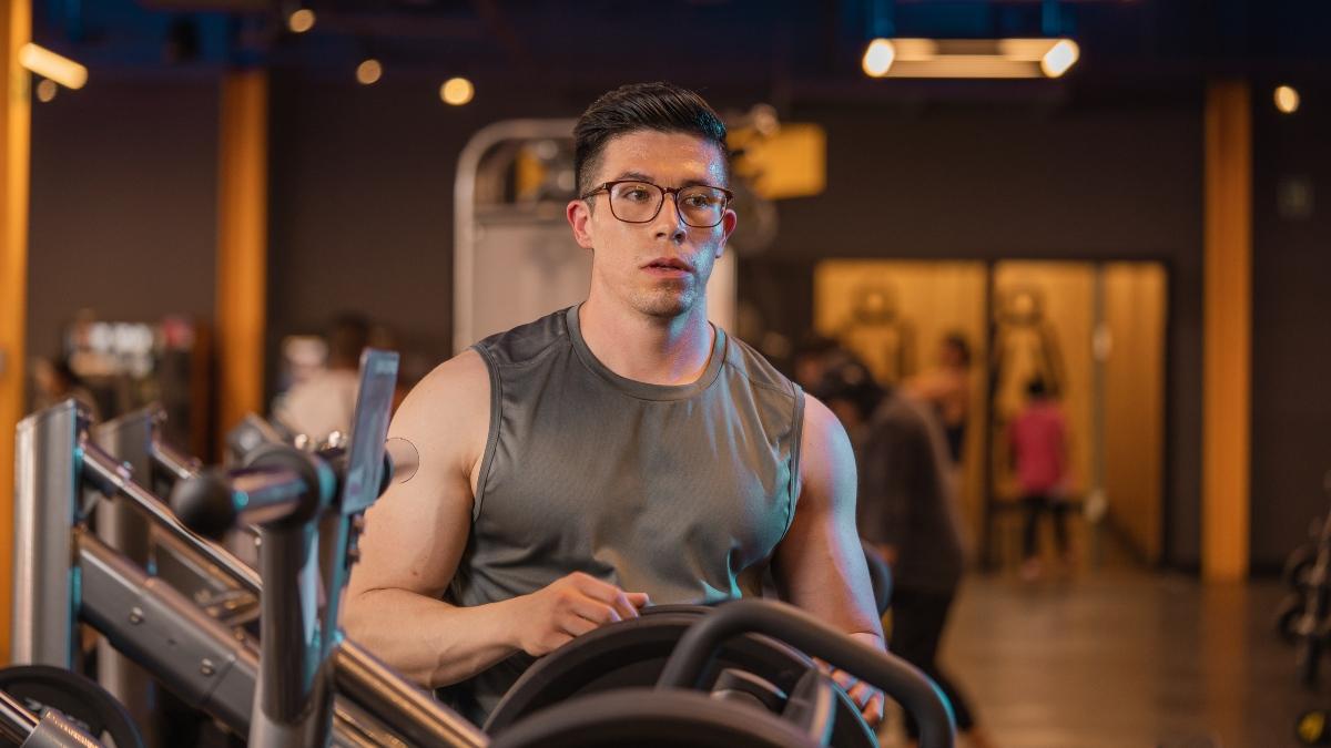 Hombre con lentes, vistiendo una playera sin mangas. En las manos sostiene un disco con el que agregará peso a una máquina del gym.