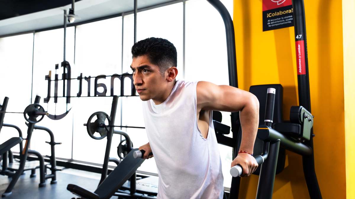 Hombre con playera sin mangas apoyando sus manos en la máquina del gimnasios para hacer fondos en paralelas