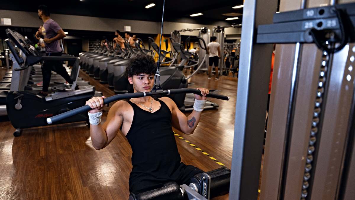 hombre sentado en una máquina de polea del gym Smart Fit. El torso inclinado hacia atrás, los brazos abiertos y flexionados. La manos están sujentado una barra.