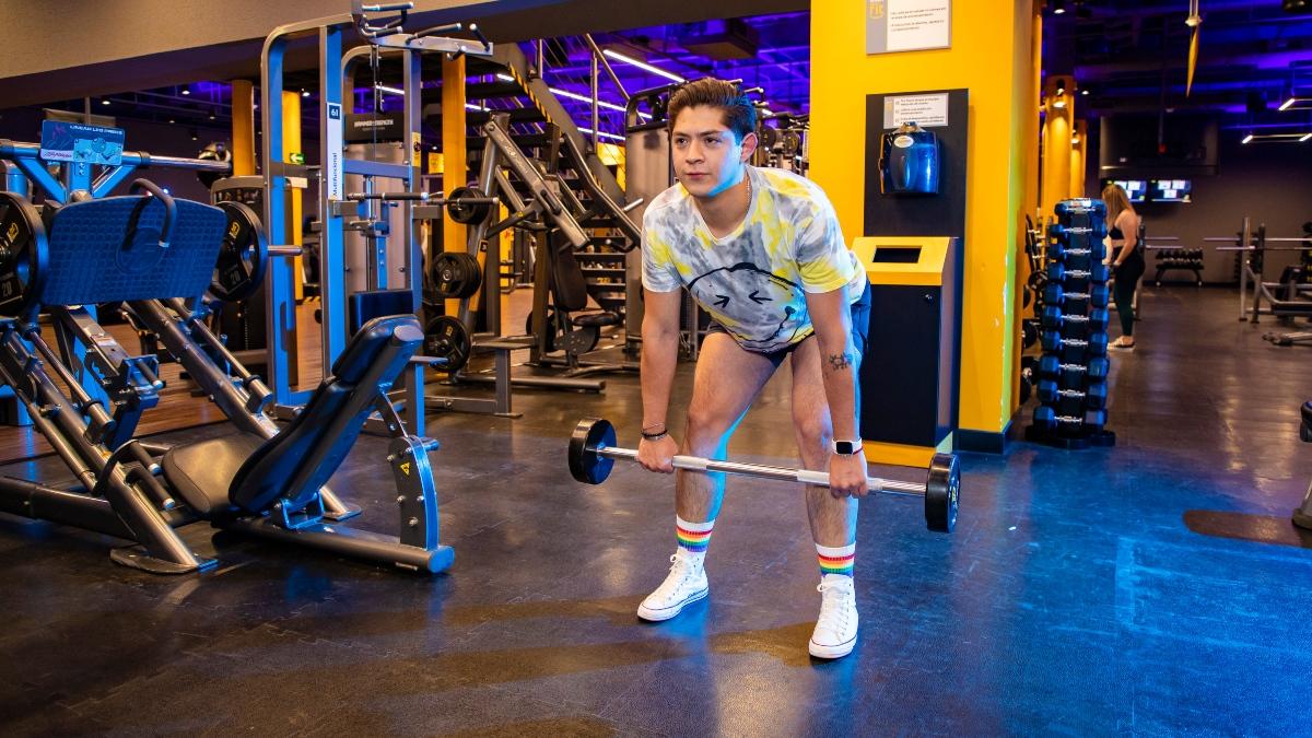 joven de pie con las piernas un poco felxionadas y el torso inclinado hacia adelante. Los brazo extendidos hacia abajo sosteniendo una barra con las dos manos, La mirada hacia el frente. Posición del remo con barra.