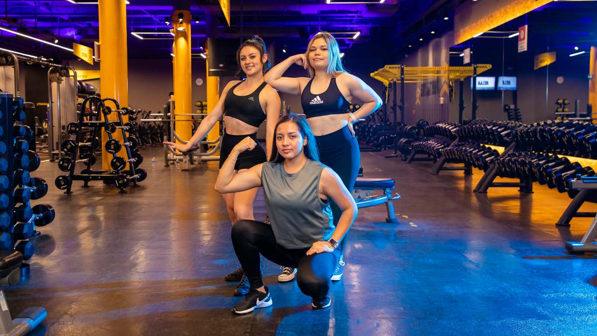 tres mujeres de frente con ropa deportiva. Una de ellas hincada, con un brazo flexionado mostrando su bicpes, el otro hacia abajo con la mano apoyada en la pierna. A su espalda dos mujeres de pie con el brazo flexionado mostrando sus bíceps.