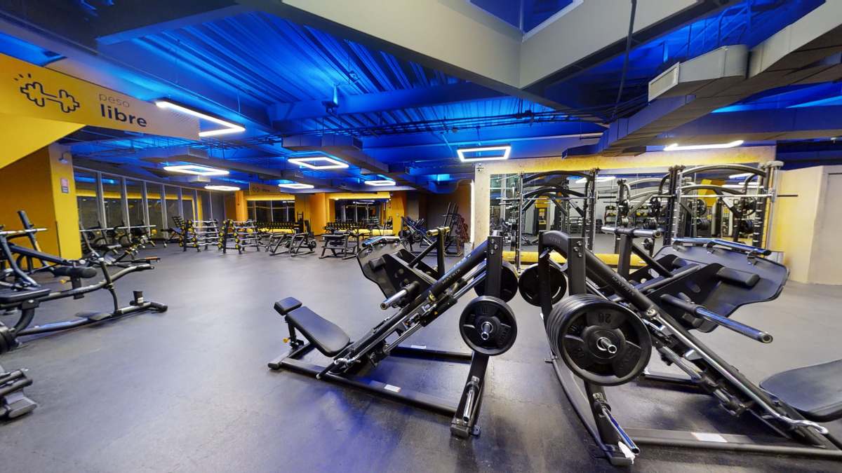 vista del area de peso libre de un gimnasio smart fit. Con una prensa para pierna en primer plano. al fondo racks con barras y mancuernas