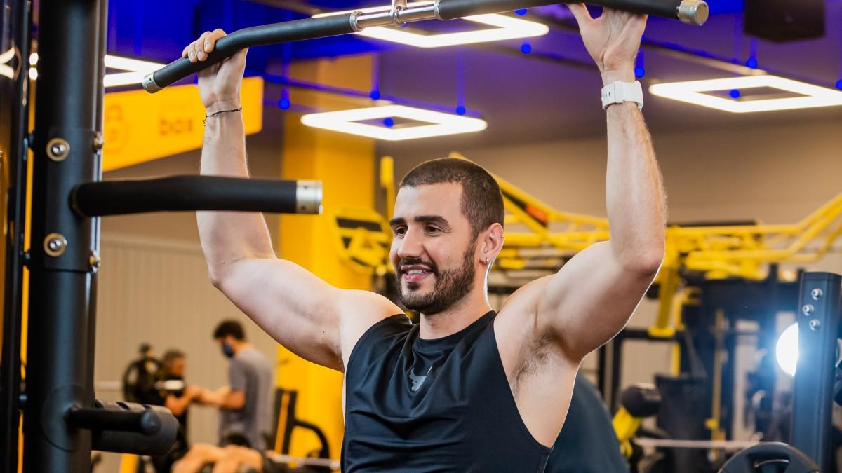 torso de un hombre sentado en una máquina de polea con los brazos levantados, tomando con las manos la barra de la polea