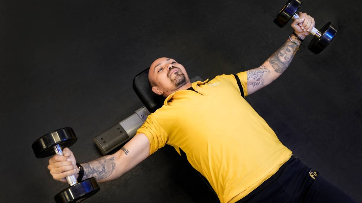 vista en contrapicada de un hombro recastado en un banco plano del gimnasio con dos mancuernas en las manos, realizando un cristo con mancuernas