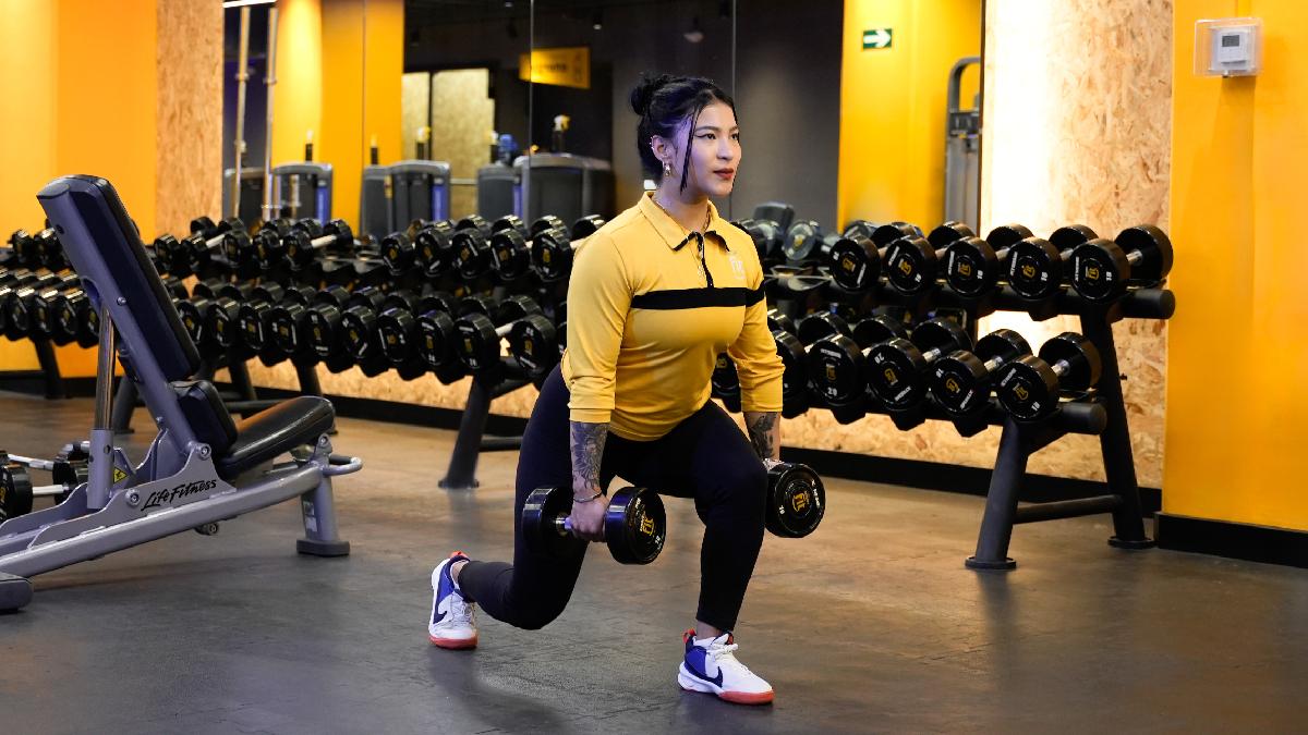 mujer con una pierna adelante y otra atrás con las rodillas flexionadas. sosteniendo una mancuerna con cada mano