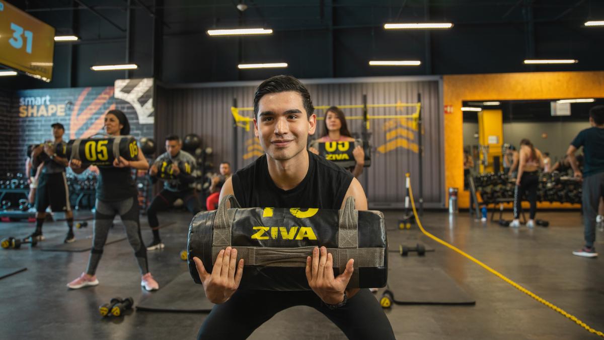 vista frontal de un hombre con las piernas flexionadas, realizando una sentadilla con un costal de arena sostenido en los brazos