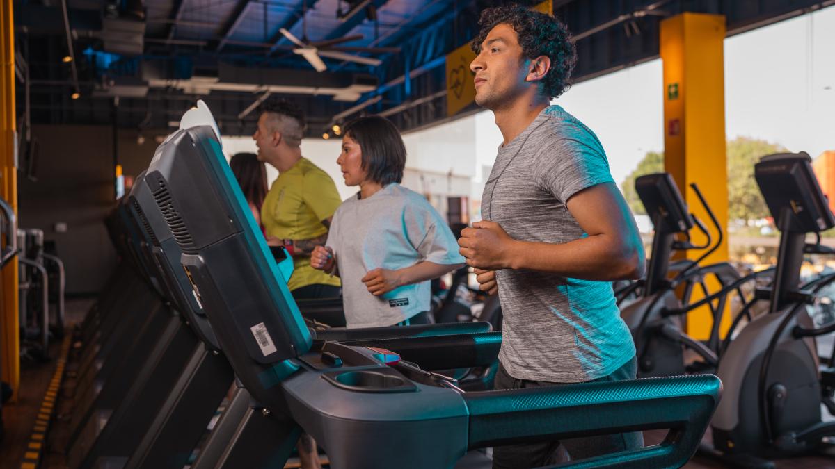 hombre en primer plano, de perfil, corriendo en una caminadora de un gimnasio smart fit. al fondo una mujer y otro hombre corriendo.