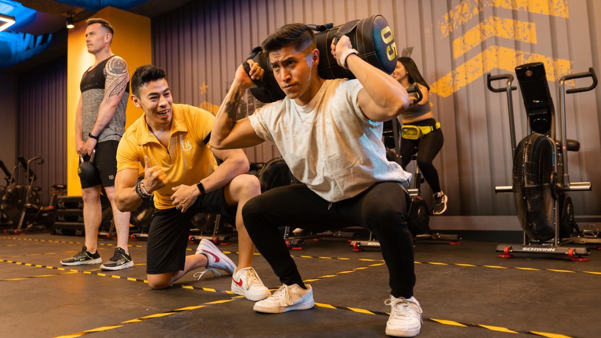hombre con las rodillas flexionadas en posición de una sentadilla con un saco de arena en la espalda, sostenido con las dos mano. a un costado un entrenador motivando, con una rodilla en el suela y la otra flexionada