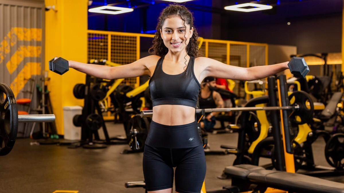 mujer de frente, de pie con los brazos extendidos a los costados sosteniendo una mancuerna con cada mano.
