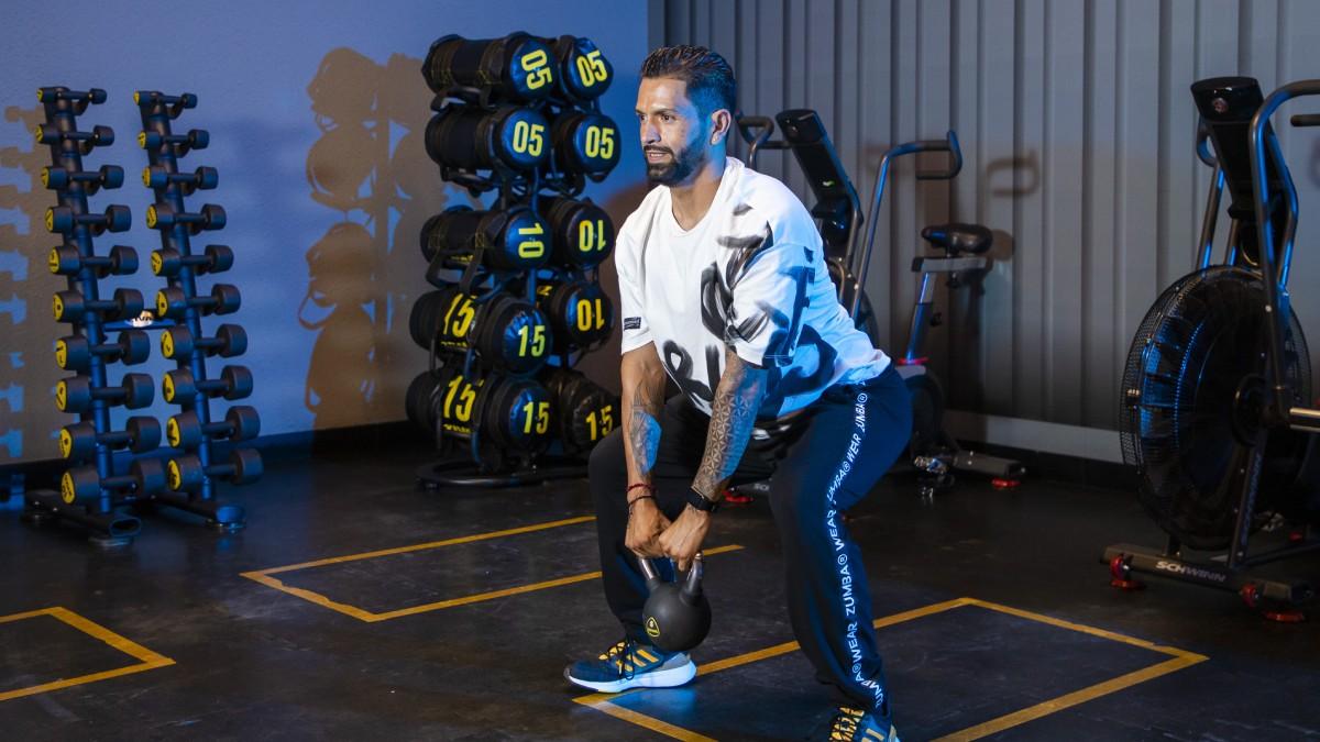 hombre de perfil con las piernas semiflexionadas en posición de sentadilla con una pesa rusa sostenida con las dos manos a la altura de las rodillas