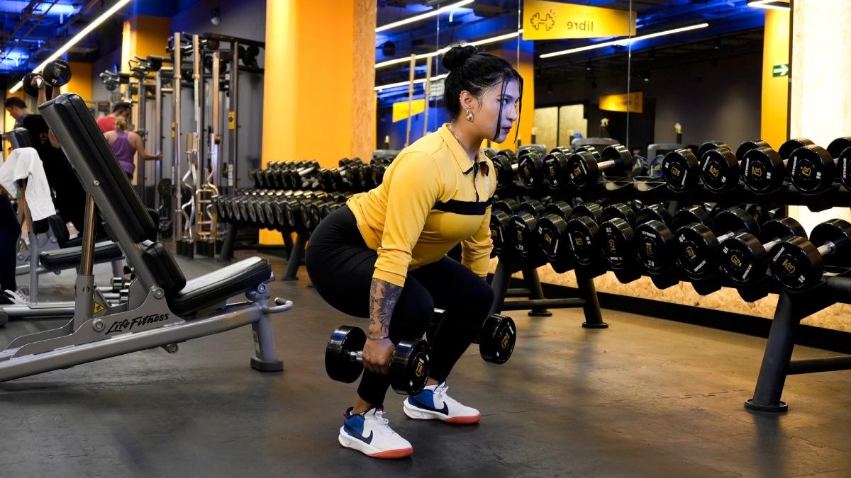 mujer de perfi, con las piernas flexionadas en posición de sentadilla con una mancuerna en cada mano