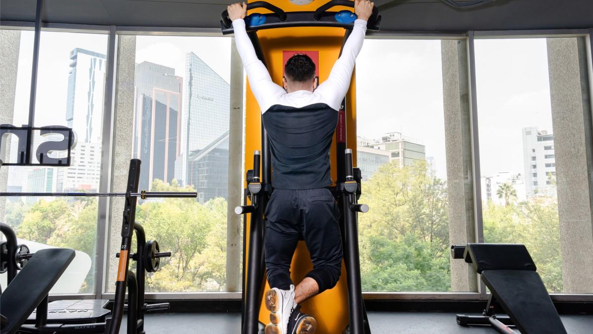 hombre de espaldas, sosteniendose con ambas manos de una máquina para hacer dominadas, con los brazos estirados y los pies cruzados