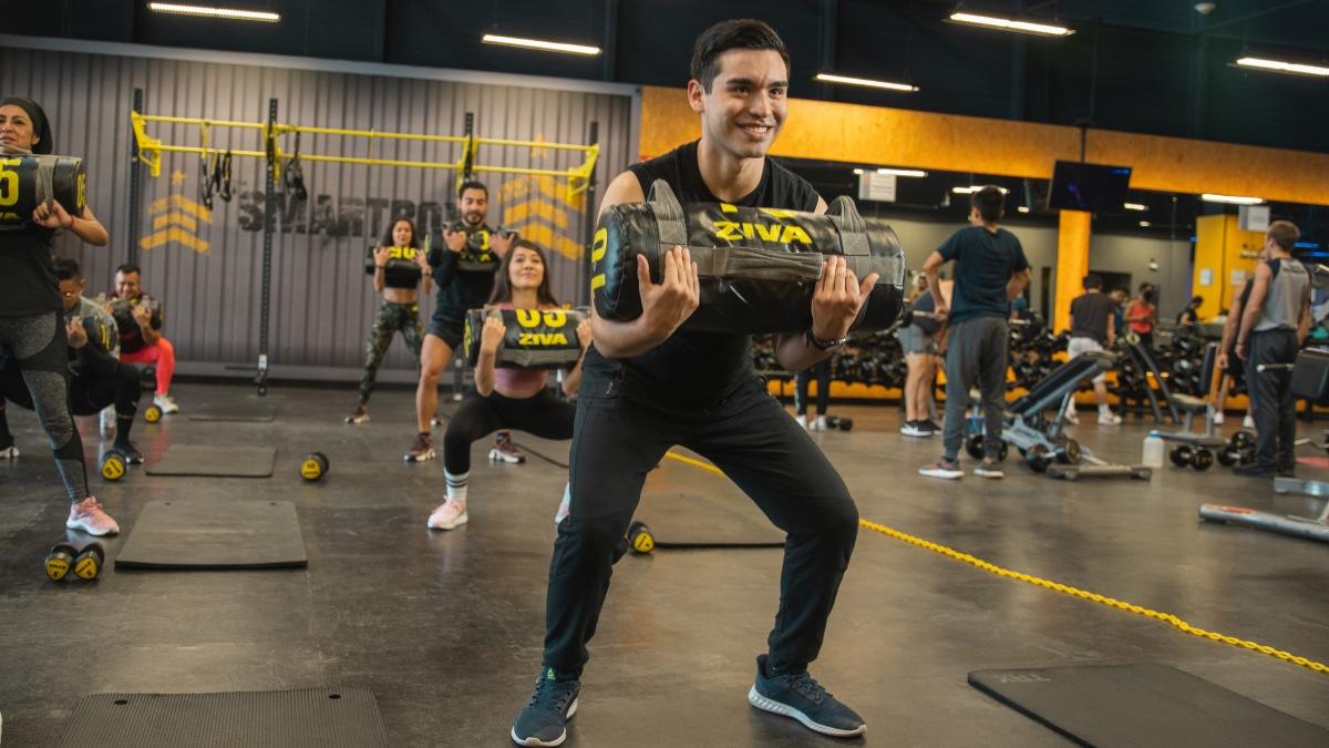 hombre de pie con las rodillas semi flexionada en posición de sentadilla con un costal de peso sostenido con los dos brazos y una sonrisa en su rostro