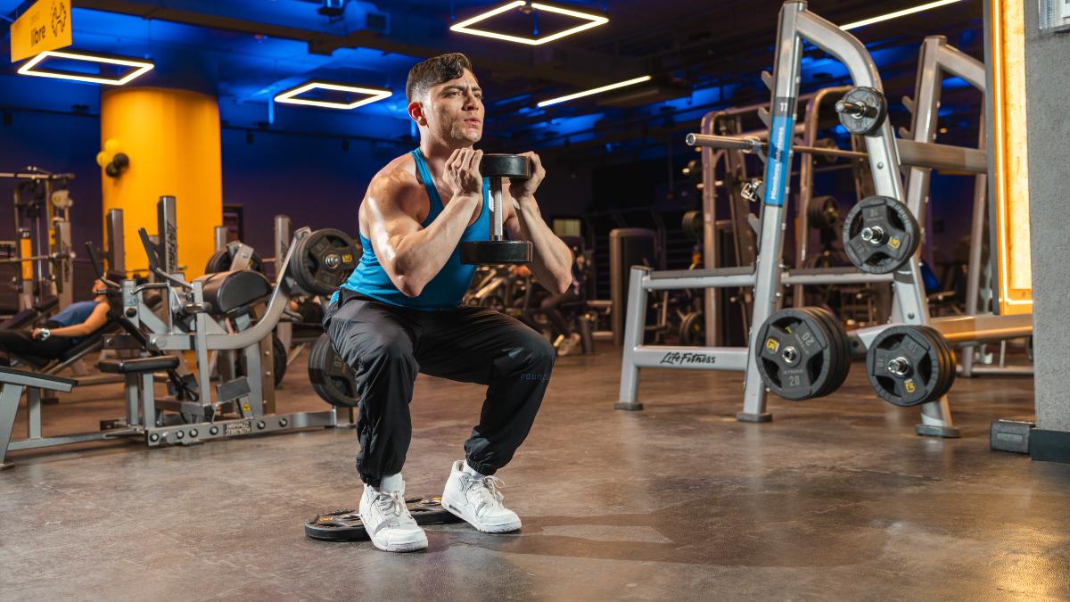 Hombre realizando sentadillas con mancuernas en gimnasios Smart Fit, enfocandose en el desarrollo de fuerza muscular en un entorno moderno y equipado.