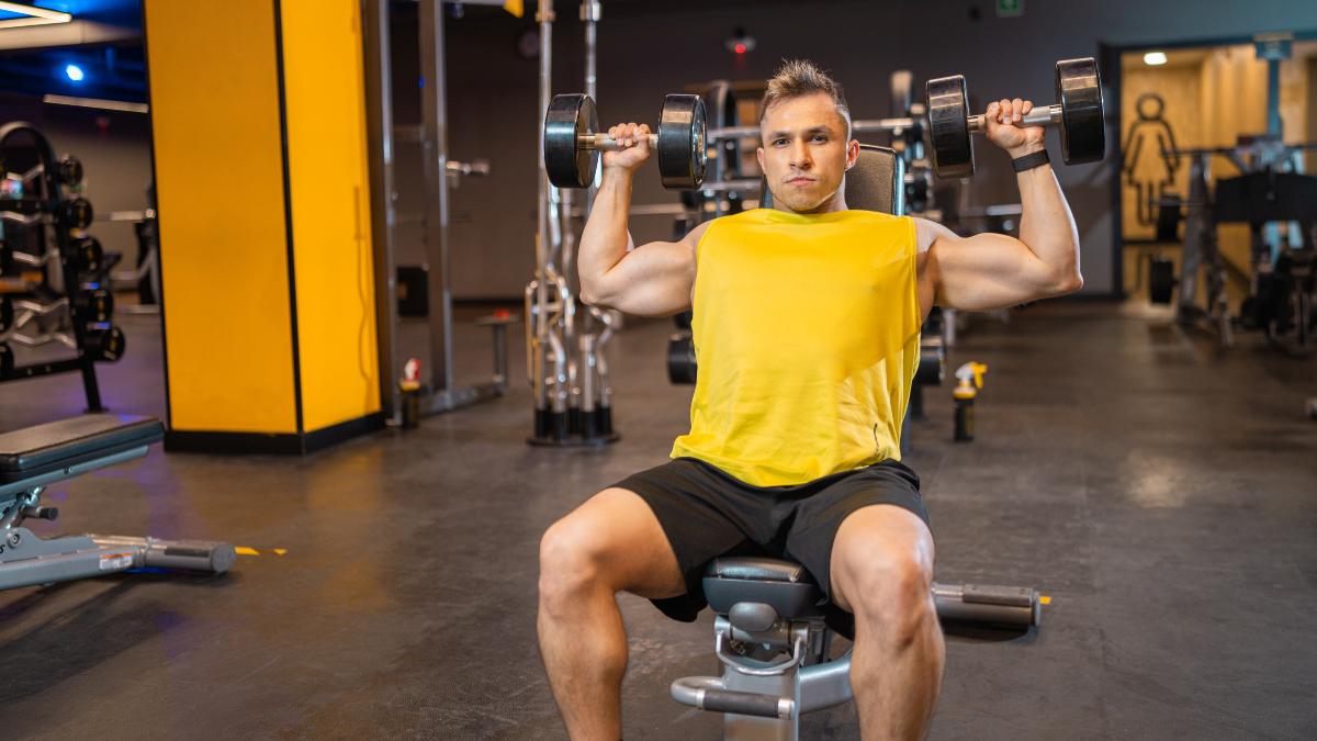 Hombre realizando press militar con mancuernas en gimnasios Smart Fit, enfocado en fortalecer los hombros en un entorno moderno y equipado.