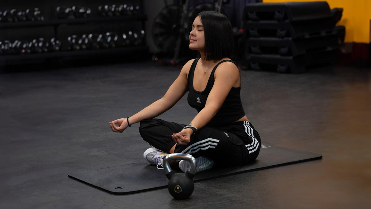 mujer sentada en el suelo en posición de flor de loto, con una pesa rusa a un lado