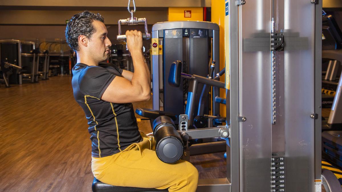 hombre de perfil, sentado en una máquina de polea en el gimnasio con las manos sujetando el triangulo del aparato