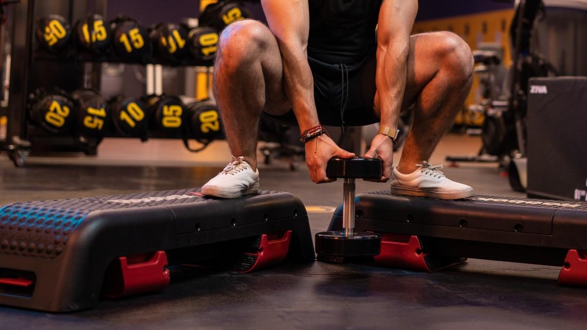torso de un hombre con los pies apoyados en dos bancos step, con las piernas flexionadas y ambas manos sosteniendo una mancuerna