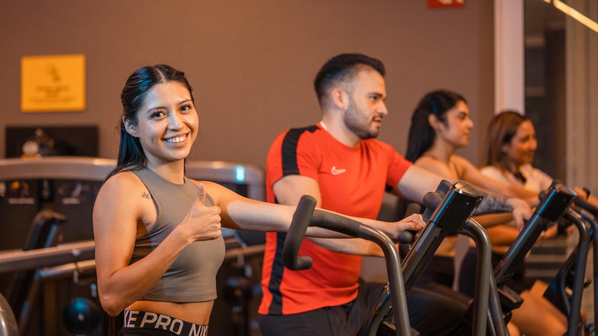 torso de una mujer en primer plano, de perfil, parada sobre una máquina elíptica del gimnasio smart fit. al fondo un hombre y dos mujeres más en las elípticas