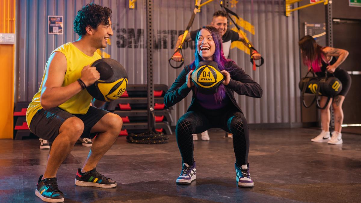 hombre y mujer de pie, sonriendo, con las piernas un poco separadas y las rodillas flexionadas, en posición baja de sentadilla. en las manos sostienen un balón medicinal a la altura del pecho