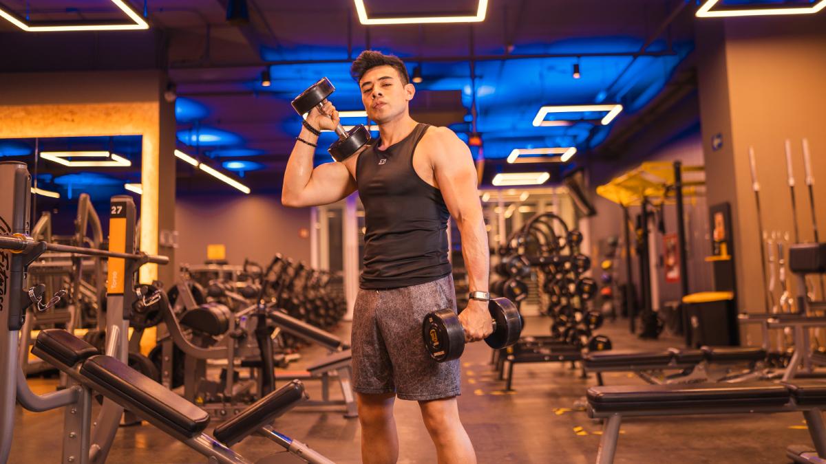 hombre de pie en primer plano. un brazo estirado hacia abajo sosteniendo una mancuerna. El otro brazo flexionado a la altura del pecho con un mancuerna en la mano