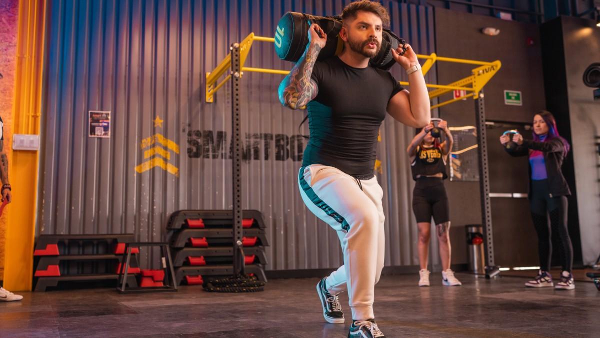 hombre de pie realizando un ejercicio de desplante con un costal de arena en los hombros, en la zona smart box de un gimnasio smart fit