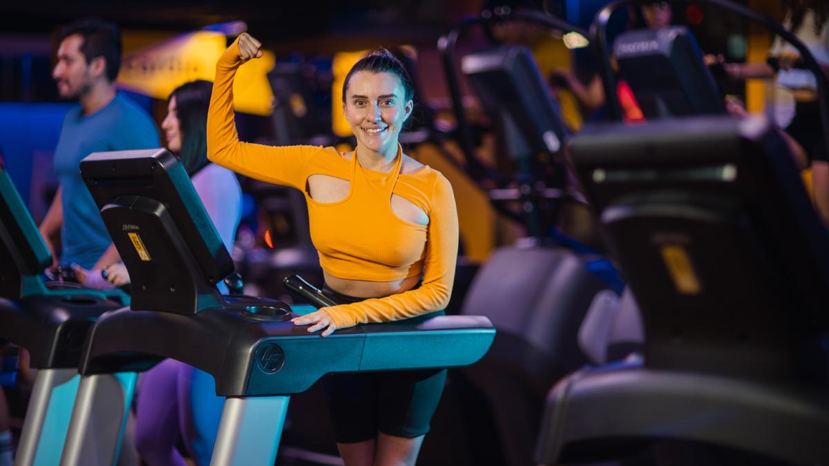 mujer parada sobre una caminadora de un gimnasio smart fit con una mano sobre la máquina y el brazo contario levantado en senal de fuerza, con el codo flexionado