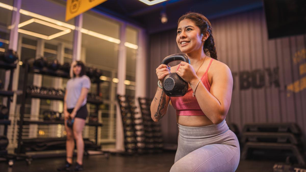mujer en posicion de desplante, con una pierna al frente felxionada y la pierna de atras flexionada con la rodilla casi tocando el suelo. sosteniendo una pesa rusa con la dos manos a la altura del pecho