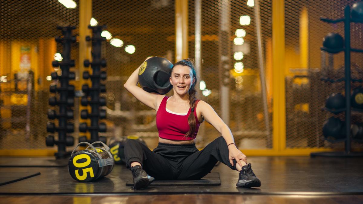 mujer sentada en el suelo de un gimnasios smart fot con las rodillas semi flexionadas, con un balon medicinal sobre un hombro sujetado con la mano y la otra mano sobre su tobillo